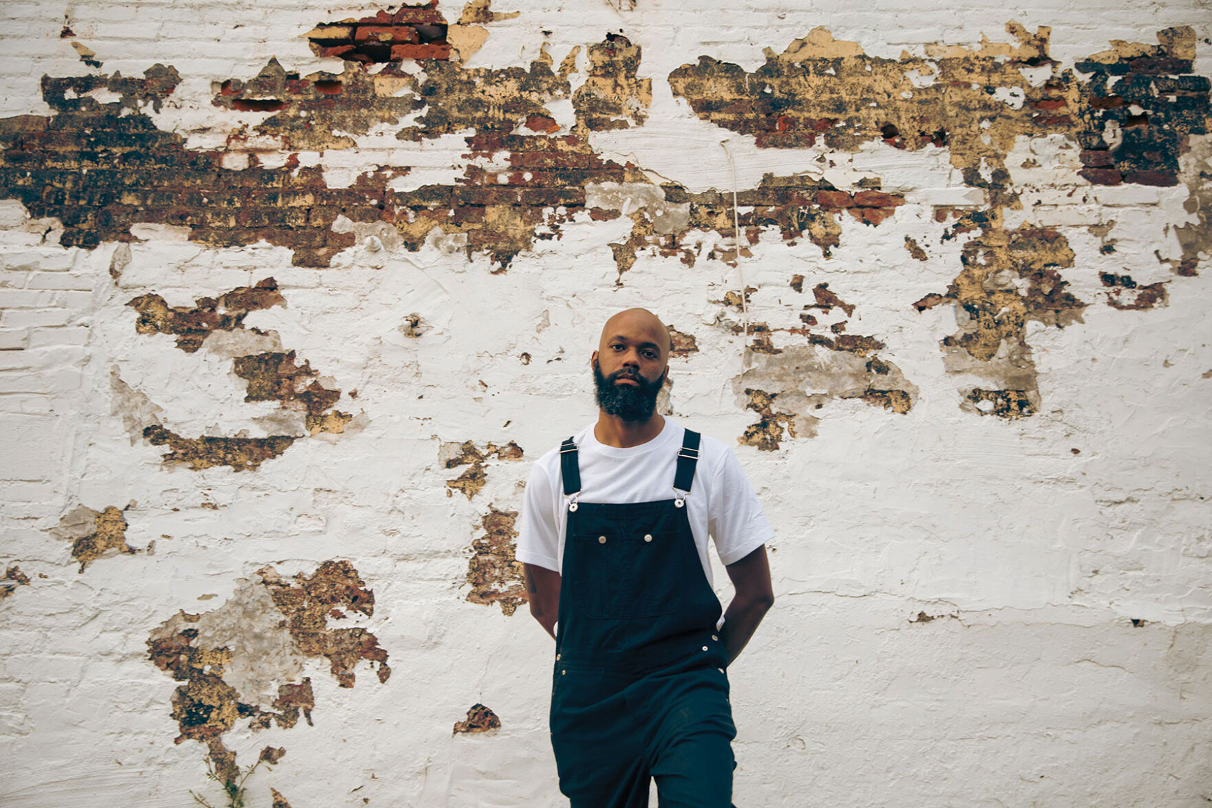 Buffalo Nichols press photo artist stands in front of a brick wall with plaster falling off